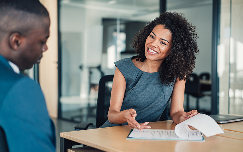 A woman sitting across a table from a man. She's smiling and pointing at a clipboard.