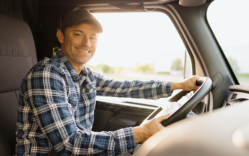 A truck driver wearing a plaid shirt and a cap driving a vehicle, holding the steering wheel with both hands.