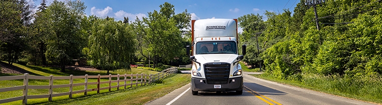 A Schneider company truck, seen from the front, driving down a two-lane county road with a wooden estate fence on one side