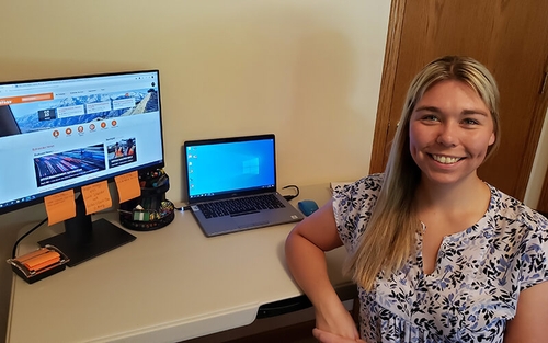 Megan Glaeser sits at her desk in her home office. On her desk is a laptop, computer mouse, pen organizer, sticky note dispenser and desktop with orange sticky notes stuck to the bottom of it.