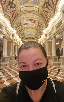 Tanaya wears a black mask and shirt and poses for a selfie in the ornate lobby of The Venetian Hotel in Las Vegas, Nevada. The lobby has a checkered floor, roman pillars and a ceiling that is covered in beautiful paintings.