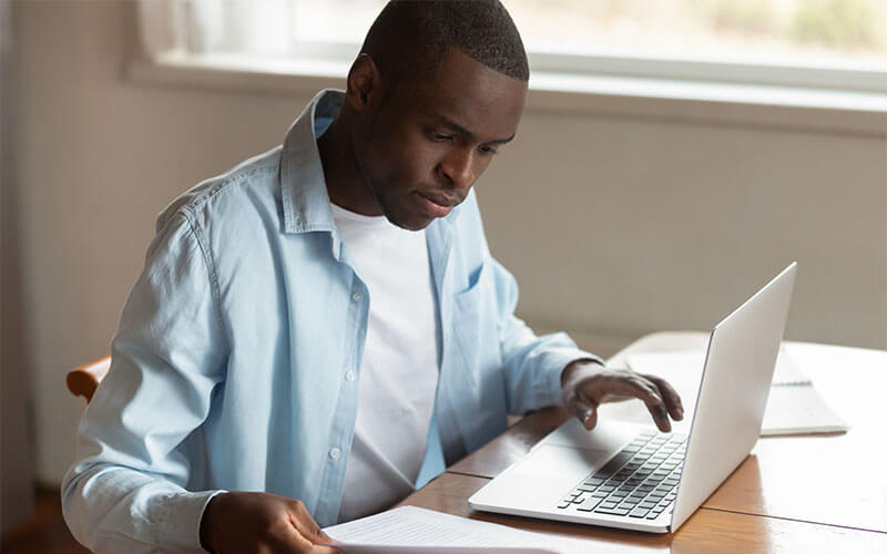 A man in a button-down shirt sits at a table and appears to be filling out tax information using paperwork and a laptop.