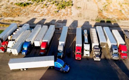 A variety of red, white, blue and silver semi-trucks hauling van trailers, flatbed trailers and tankers are parked side-by-side in semi-truck parking spaces. A blue truck hauling a white trailer appears to be lining up to back into a spot.