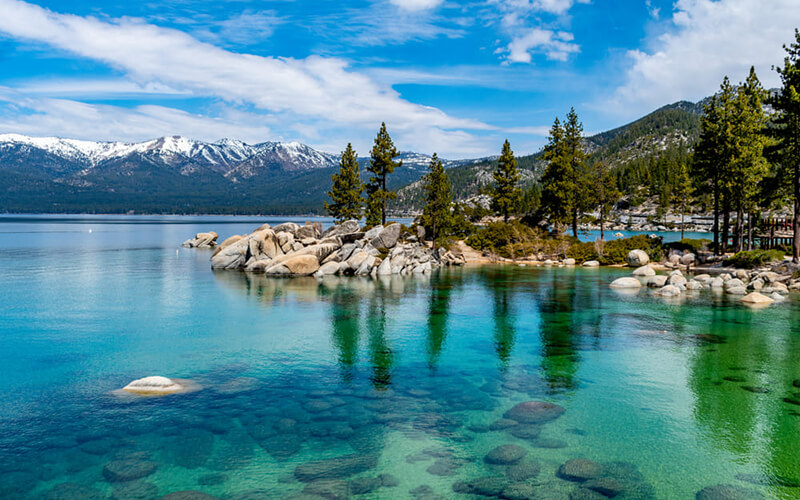 A beautiful view of Lake Tahoe includes clear emerald water, an outcropping of rocks with pine trees going out into the water and features mountains in the background.