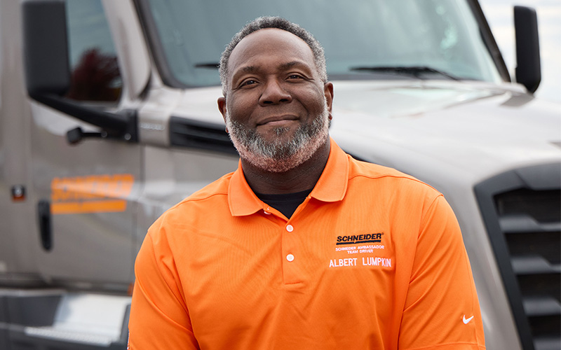 Albert Lumpkin standing outside in front of a gray Schneider semi-truck, smiling straight ahead.