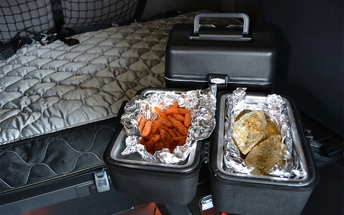 Two 12-volt portable stoves sit on a counter in a sleeper semi-truck. One portable stove is closed and the other is open and contains carrots and chicken wrapped in tin foil.