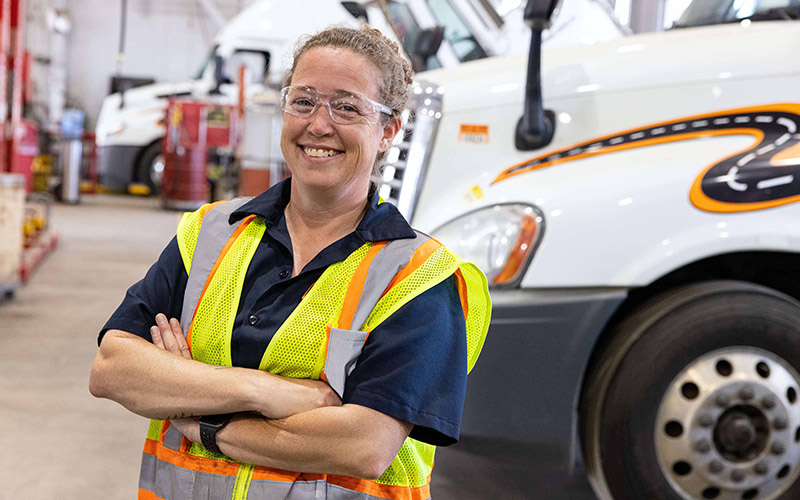 A woman standing and smiling in front of a Schneider semi-truck with her arms crossed. She's wearing a yellow safety vest and safety glasses. 