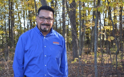 Robert Soto smiles while standing in the woods, with his hands in his pockets and wearing a Schneider button-up shirt.