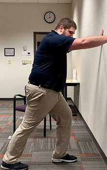A physical therapist demonstrates stretching his leg by staggering his right foot behind his left and pushing against the wall.