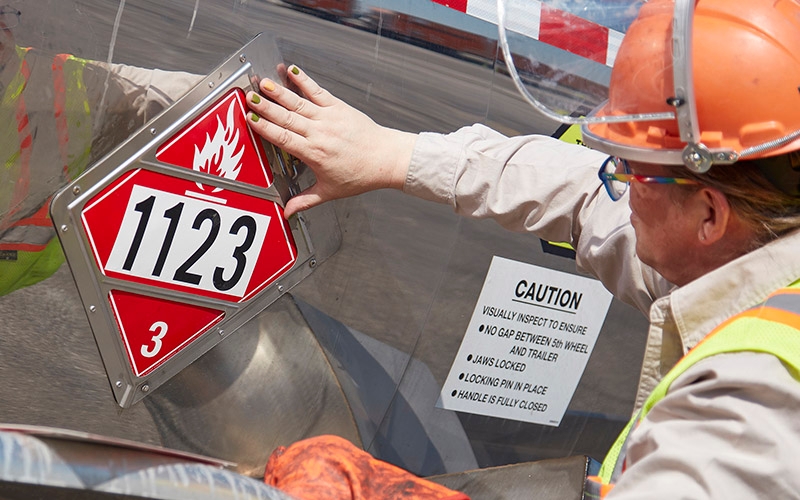 A HazMat truck driver in PPE inspecting a red placard on the side of their tanker trailer.