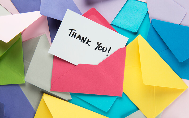 Envelopes varying in color lay in a pile. The middle most envelope is opened and inside is a white note with the text, "Thank you!"