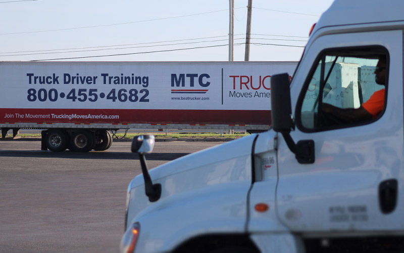 The cab of a white semi-truck in front of a separate white trailer that reads "truck driver training" in the background.