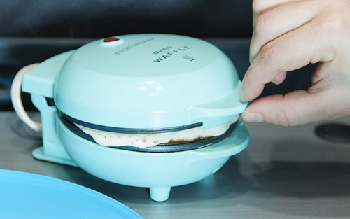 A light blue mini waffle maker with a waffle cooking inside it. A person’s hand is visible, reaching towards the mini waffle maker, possibly to check the cooking progress or open it.