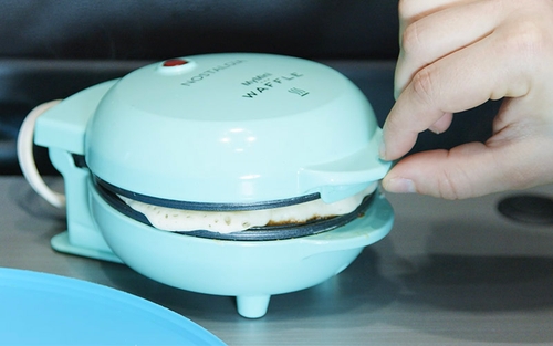 A light blue mini waffle maker with a waffle cooking inside it. A person’s hand is visible, reaching towards the mini waffle maker, possibly to check the cooking progress or open it. 