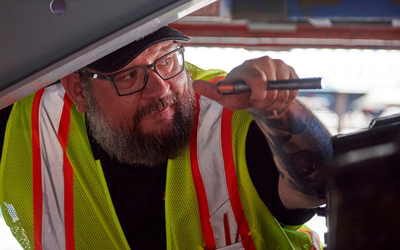 A truck driver in a high-visibility vest inspecting a semi-truck with a flashlight in an industrial setting.