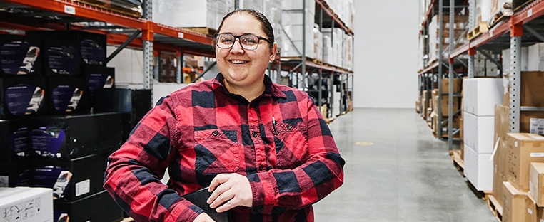 A Schneider warehouse associate stands in a row it wall shelves with pallets of boxes.