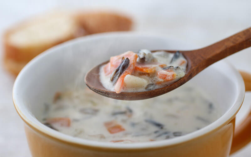 A wooden spoon full of steaming chicken wild rice soup is held directly over an orange cup of soup with a white interior.