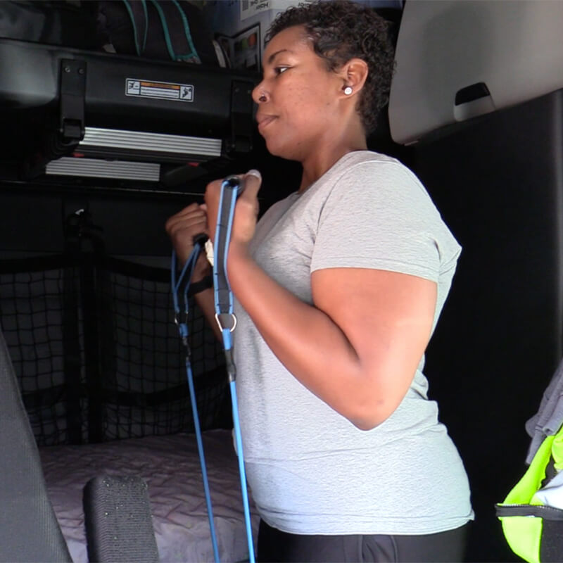 A female team driver performs bicep curls using a resistance band in the cab of her truck.