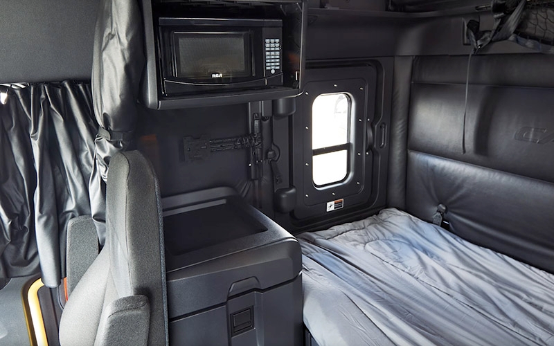 The interior of a truck’s sleeper berth. It features a bed with a grey blanket, black walls and a small window with curtains. A microwave is mounted above the foot of the bed, and there is a dark-colored storage compartment next to the bed.