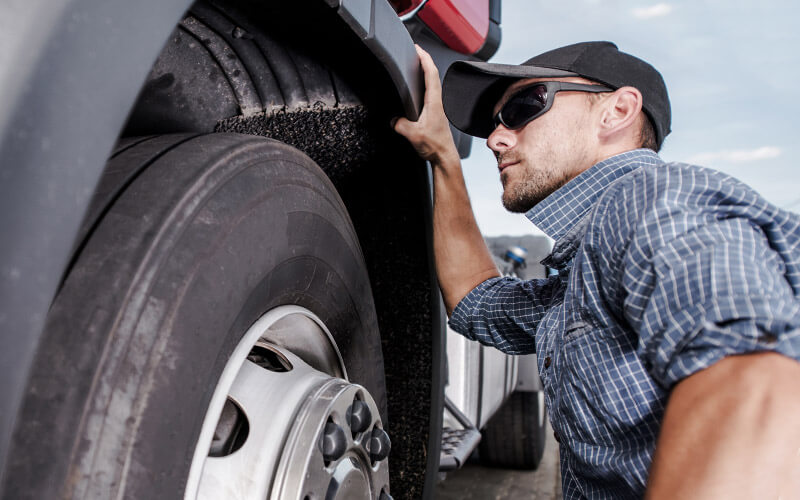 A truck driver wearing a hat and sunglasses kneels to inspect his semi-truck's wheel.