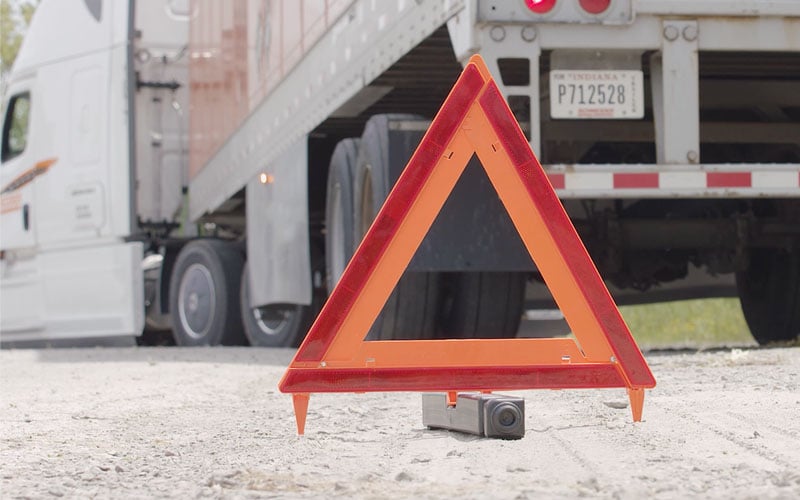 A reflective orange emergency triangle on the gravel behind a stopped semi‑truck to signal a roadside emergency.
