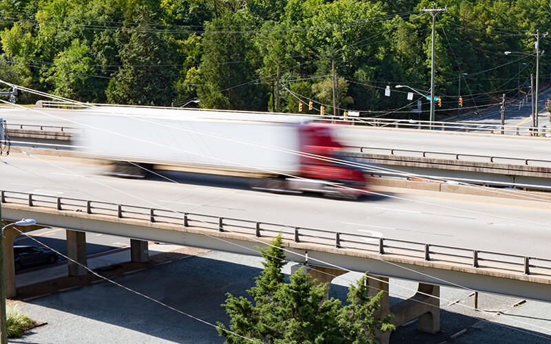 A red semi-truck hauling a white dry van container speeds by in a blur on a highway overpass.