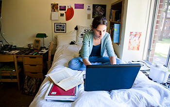 A young woman sits on her bed in a college dorm room with a laptop, book and notebook in front of her.