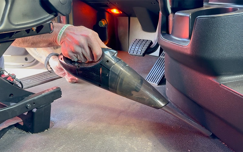 A Schneider truck driver using a handheld vacuum to clean the floor by the driver's seat of their semi-truck. 