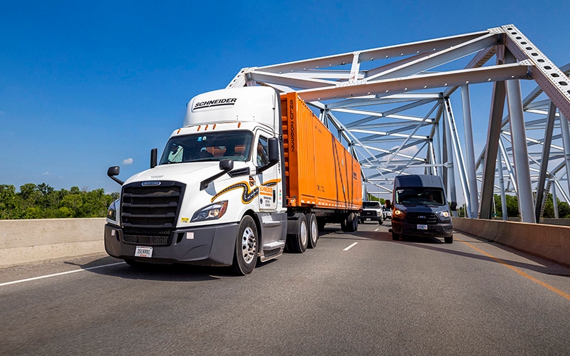 A white Schneider day-cab semi-truck hauling an orange intermodal container drives across a bridge.