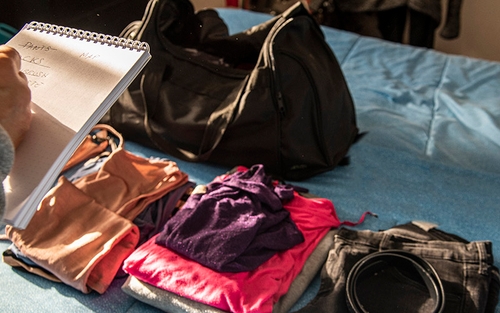 Someone holding a list stands over a bed that has a bag and clothing items laid out on it.