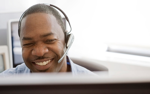 A smiling man sits in front of his computer with a headset on.