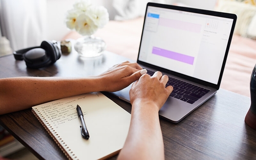 Hands type on a laptop that is sitting on a wooden desk that also has a notebook, pen, headphones and flowers on it.