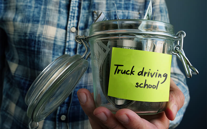 A man in a plaid shirt holds an open jar with money in it. There is a sticky note on the front that says "truck driving school."