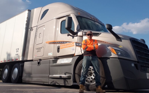 Joel Torres-Sanchez, a truck driver wearing a neon orange long sleeve shirt, blue jeans and work boots, stands proudly in front of his brand-new grey 2022 Freightliner Cascadia semi-truck.