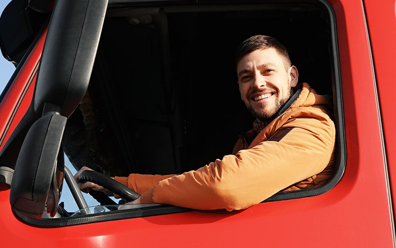 A man in an orange coat smiling while sitting in the driver's seat of a red semi-truck.