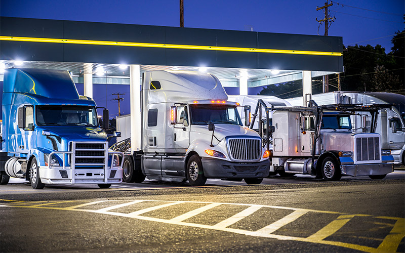 Three semi-trucks, one blue, one white and one silver, parked side by side at a brightly lit truck stop at night.