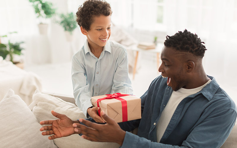 A child gives a wrapped present to an adult who is sitting on a couch.