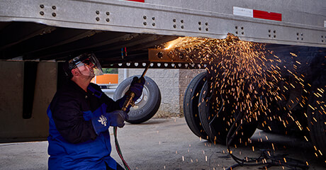A Schneider diesel technician, wearing protective gear, is using a welding torch to work on the underside of a large trailer.