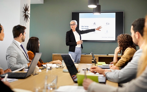 A group of people sit around a table with their laptops and watch a woman who is standing in the front of the room. The woman is pointing to a projector and talking.