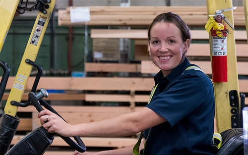 A female warehouse worker drives a forklift through a warehouse.