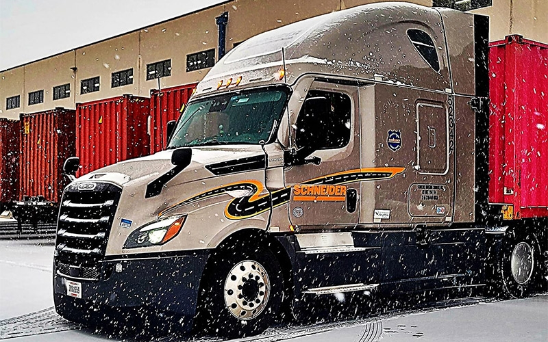 A grey Schneider semi-truck parked in a snowy lot in front of other orange trailers.