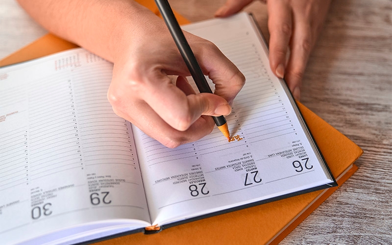 A hand using an orange market to write a list in a lined calendar.