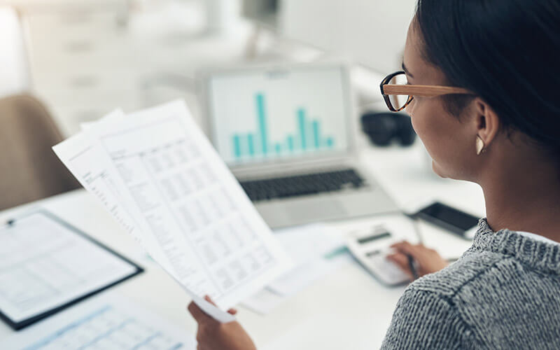 An accountant holds up a financial document and looks at it closely while tapping numbers into her calculator with her other hand.