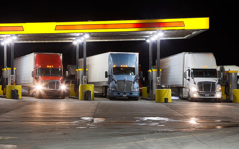 Three semi-trucks are parked next to fuel pumps at a truck stop.