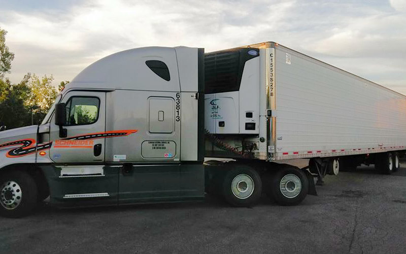A silver Schneider truck parks at an angle in a parking lot with a white refrigerated trailer attached to the semi-truck.