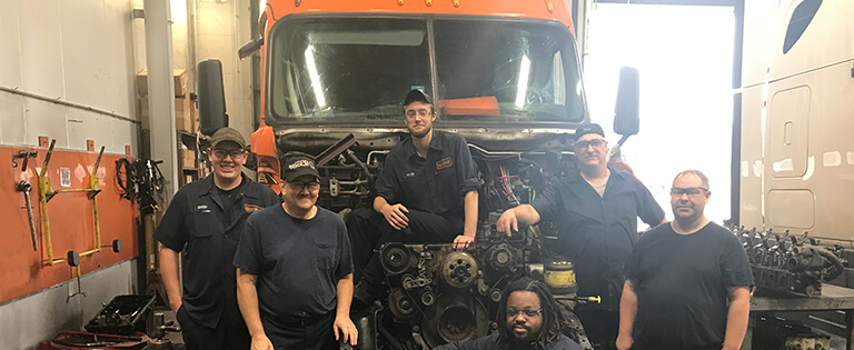 A group of six Schneider diesel technicians pose in various ways with a hoodless company truck