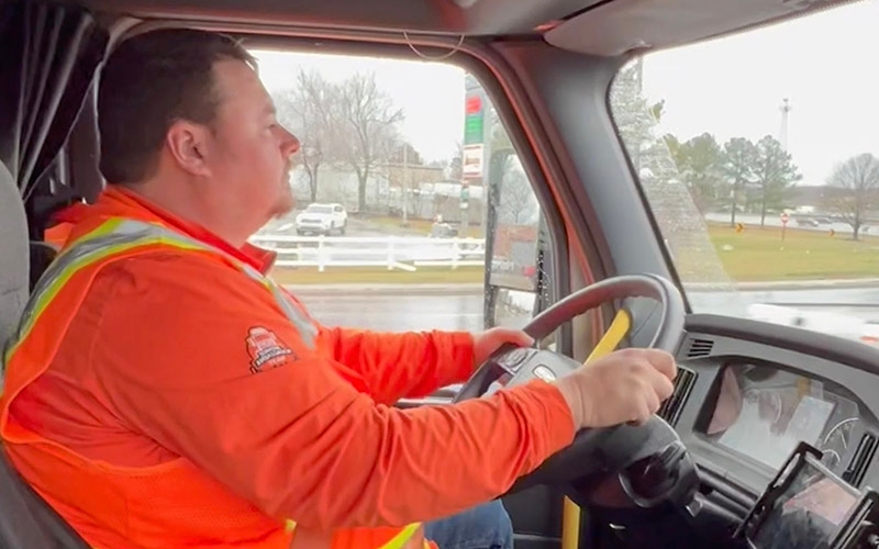 A Schneider driver in a high-visibility orange jacket and safety vest behind the wheel of a semi-truck.