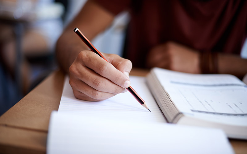 Someone wearing a red shirt sits at a table and studies a book while taking notes.