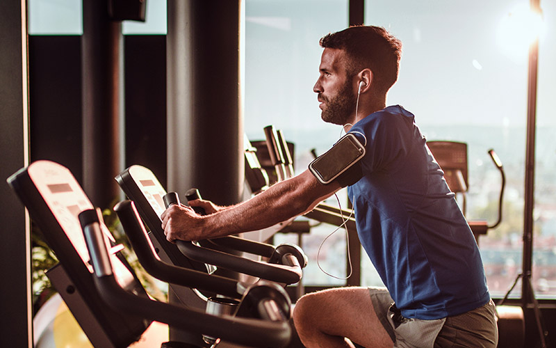 A person on an exercise bike in a gym with large windows. The person is wearing a blue shirt, black shorts and has an armband holding a phone.