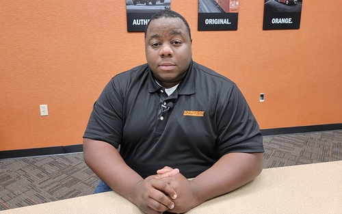 Driver Joseph Morning sits at a table and and smiles. He wears a black polo shirt and sits in front of an orange wall in a conference room.